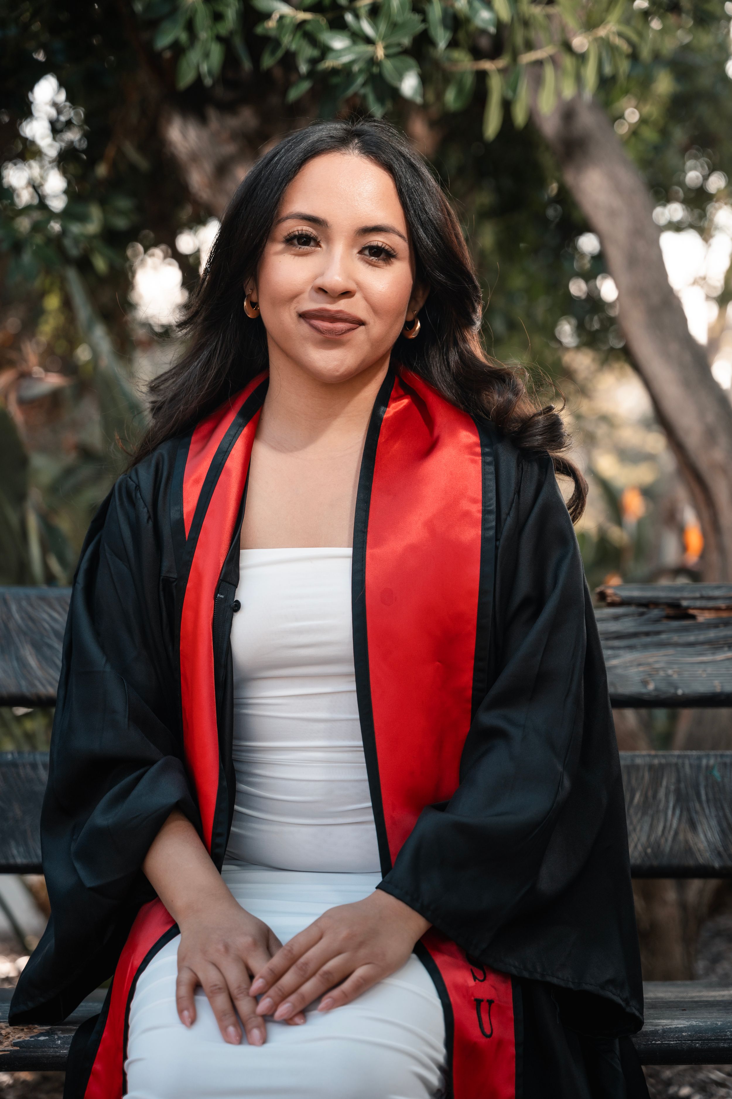 Graduate wearing traditional black graduation gown with red stole seated outdoors.
