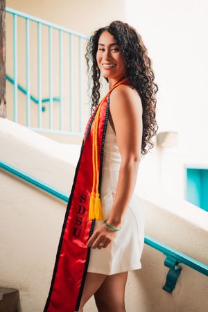 Graduate in white dress and red stole poses near turquoise railing.