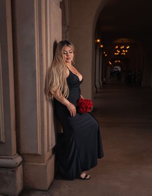 Elegant figure in black evening gown poses with red roses in dimly lit archway.