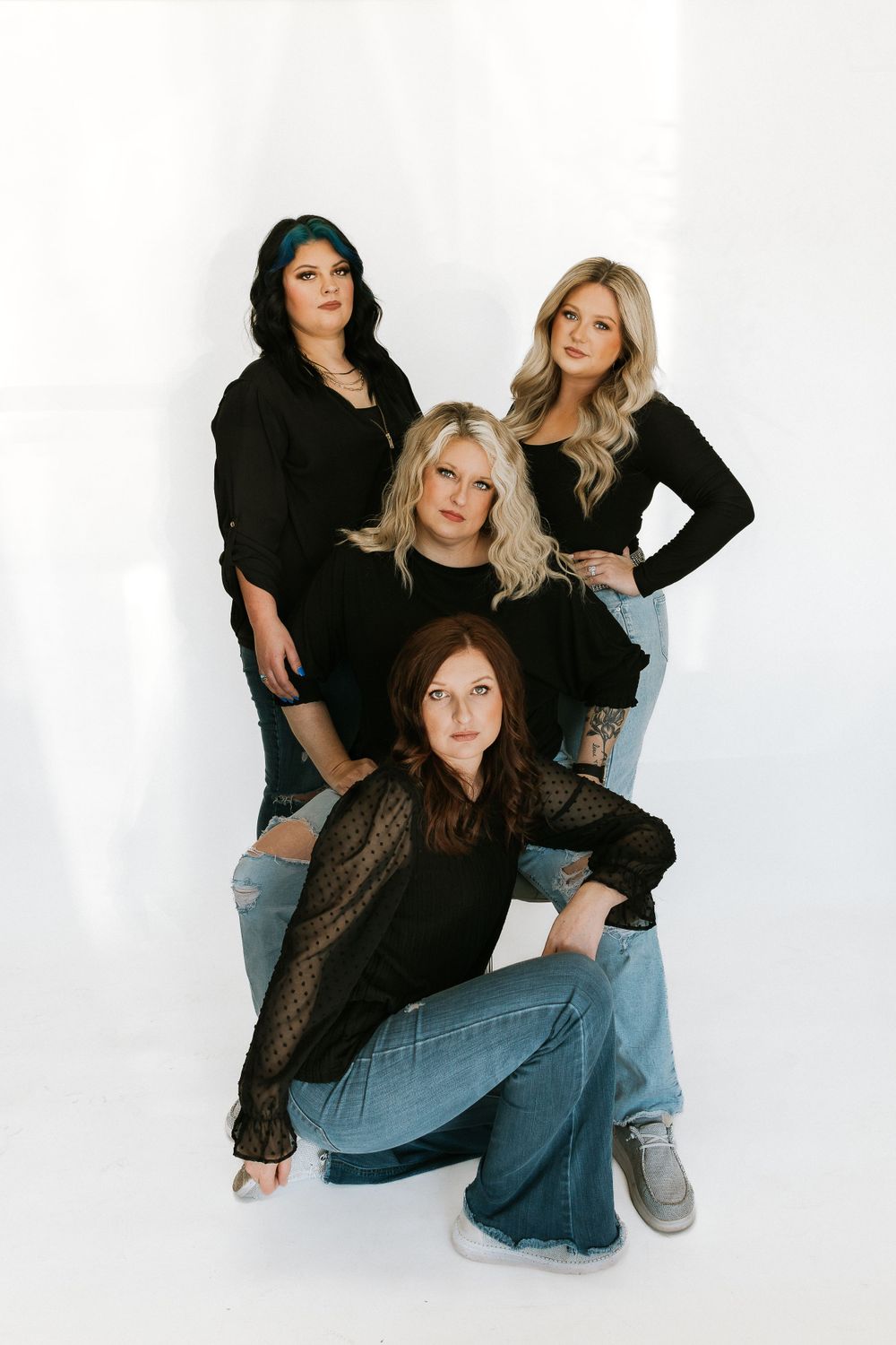 Four women in black outfits and blue jeans pose together against a white background in a fashion portrait.