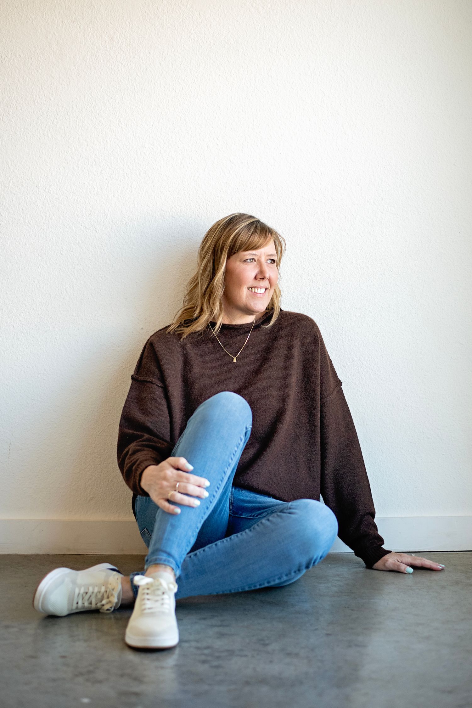 Person in brown sweater and jeans sitting casually against white wall.