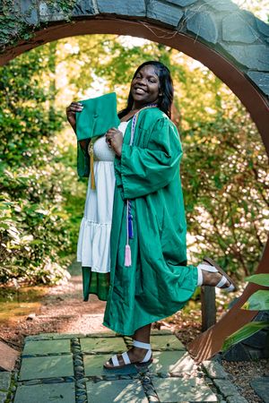Graduate in emerald green commencement gown poses joyfully under a garden archway during outdoor photo session.
