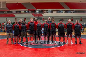 A group of athletes stands together on a red mat with a circular logo in a sports gymnasium.