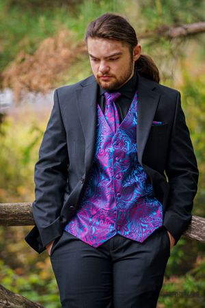 A black suit with vibrant blue and pink paisley patterned vest stands on a wooden railing in an outdoor setting.