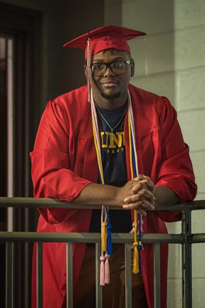 Graduate in red cap and gown with honor cords poses against railing in hallway.