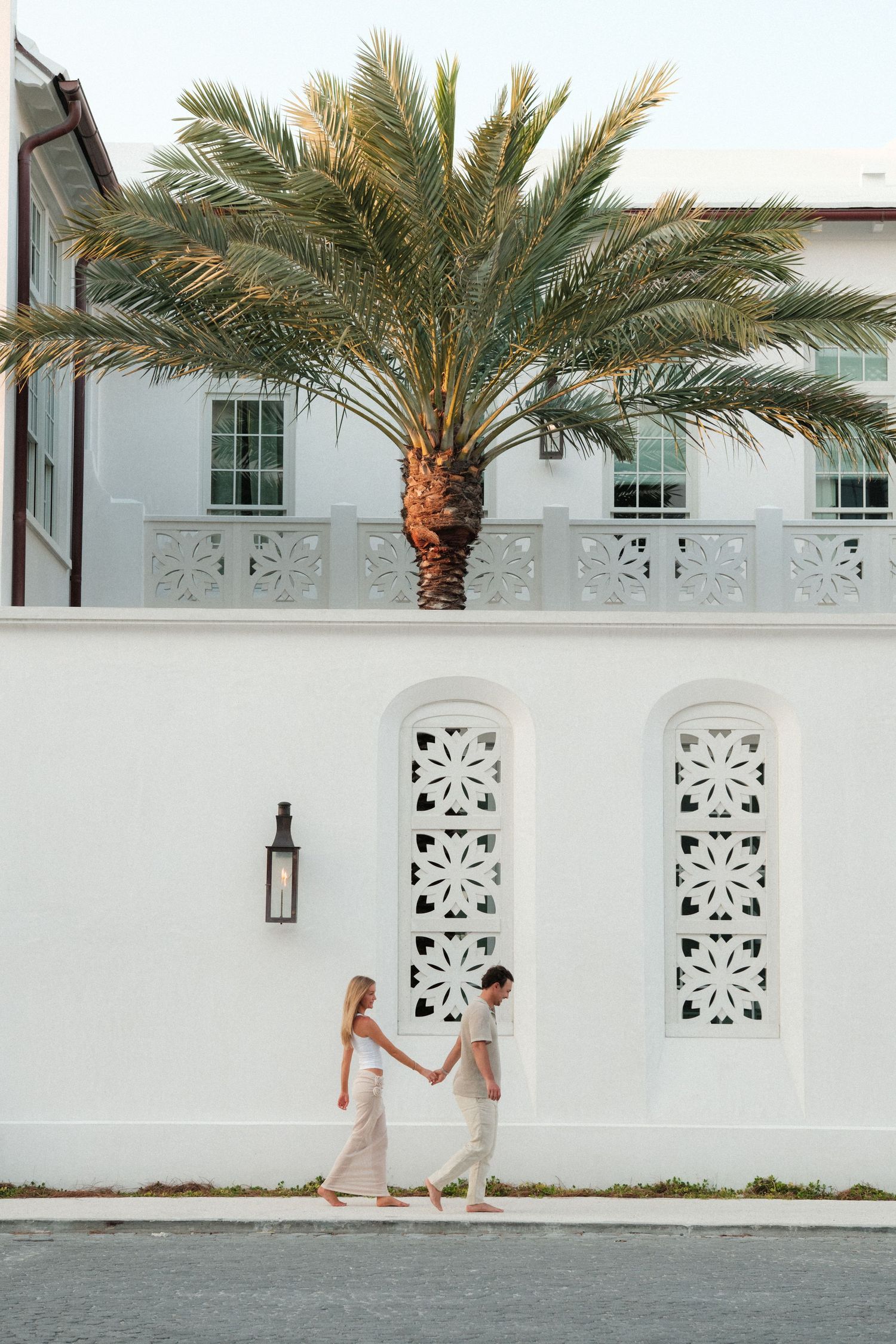 Couple walking hand in hand past a white Mediterranean-style building with ornate details and palm tree.