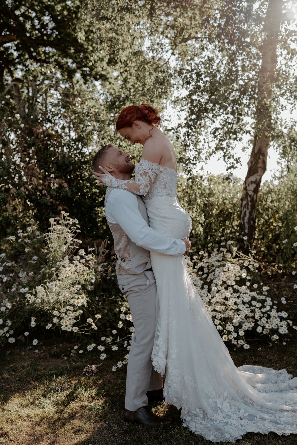 A romantic wedding couple embraces in a sunlit garden with white flowering trees creating a dreamy backdrop.