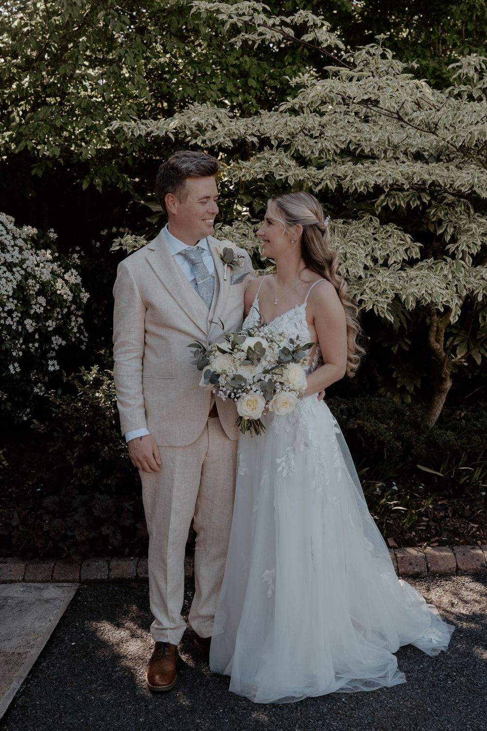 A smiling bride and groom pose together outdoors, she holds a white bouquet, he wears a beige suit, with lush greenery behind them.