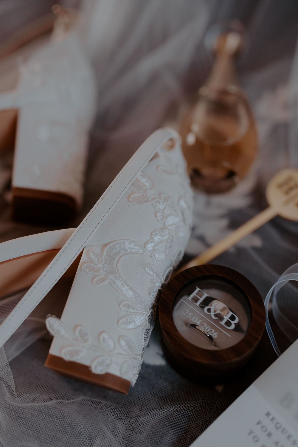Elegant wedding flat lay with lace bridal shoe, wooden ring box with H&B initials, and golden honey jar.