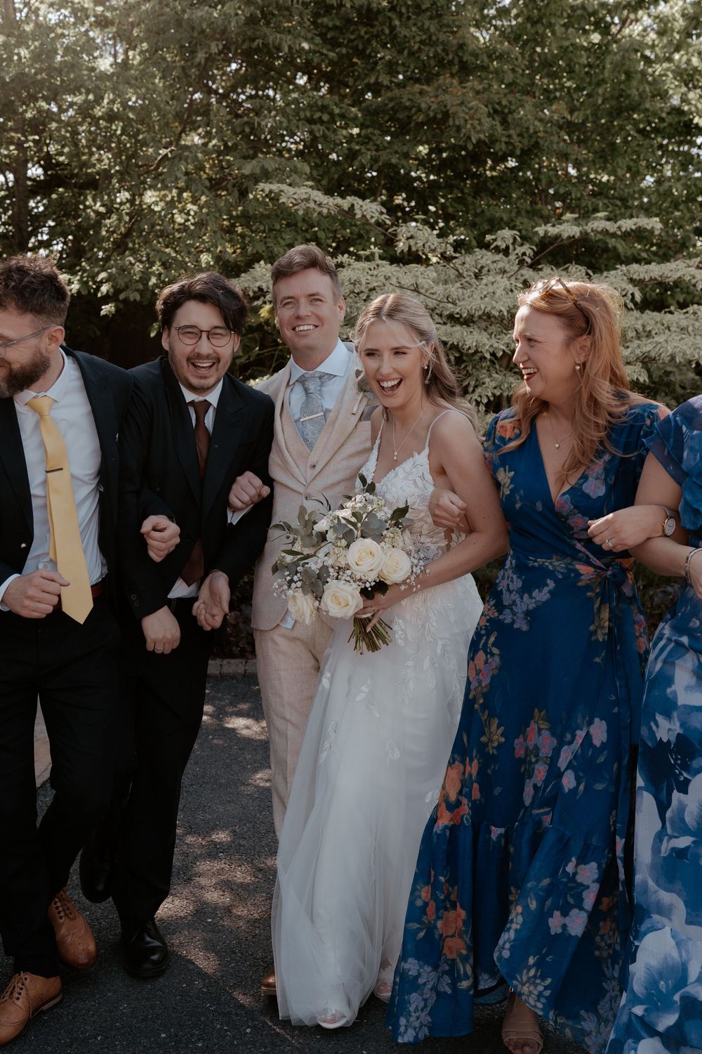 A joyful wedding party walks outdoors, bride holding white bouquet in lace gown, groom in beige suit, guests laughing.