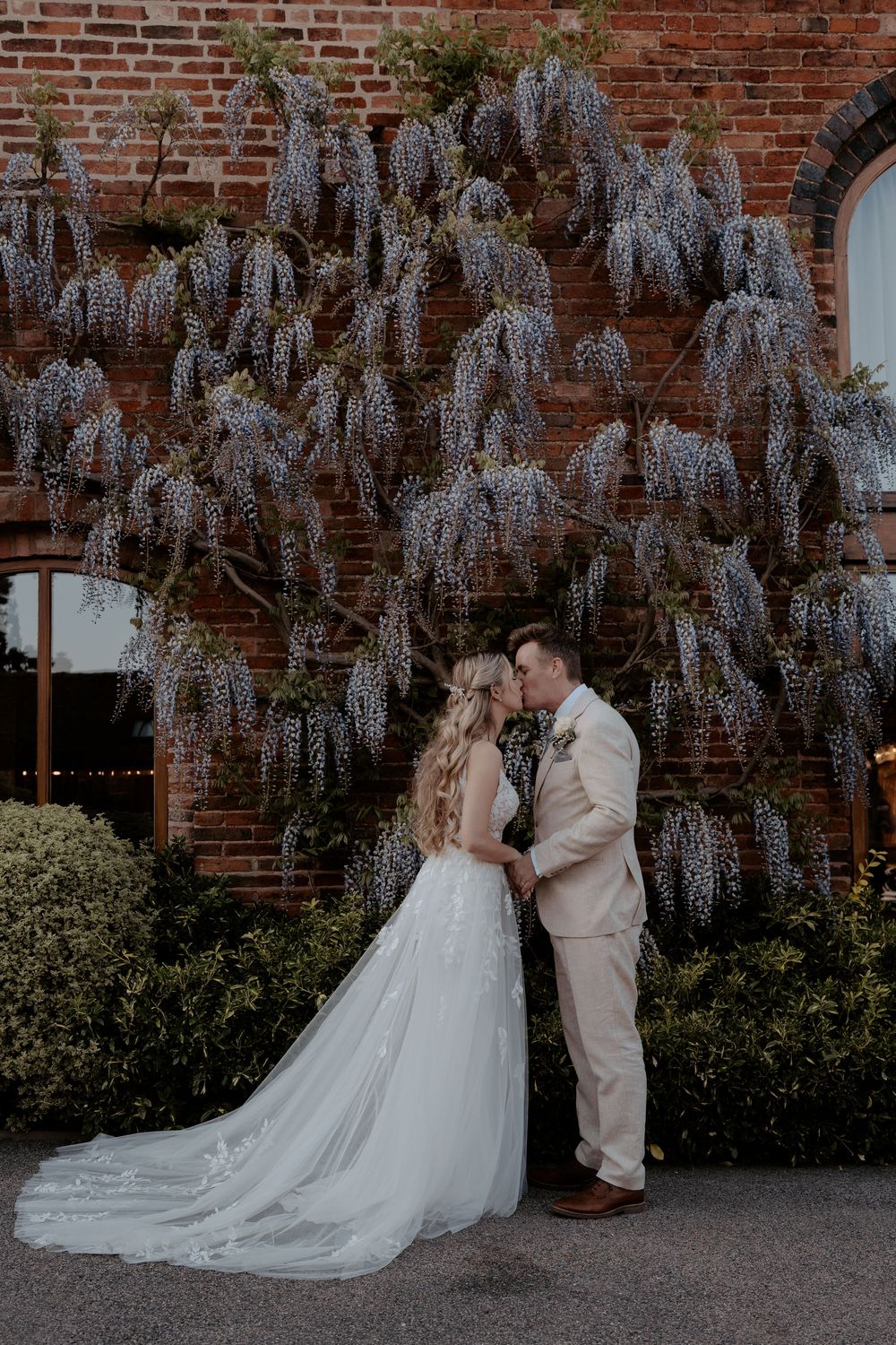 Bride and groom sharing a kiss in front of a wisteria-covered brick wall at an outdoor wedding venue.