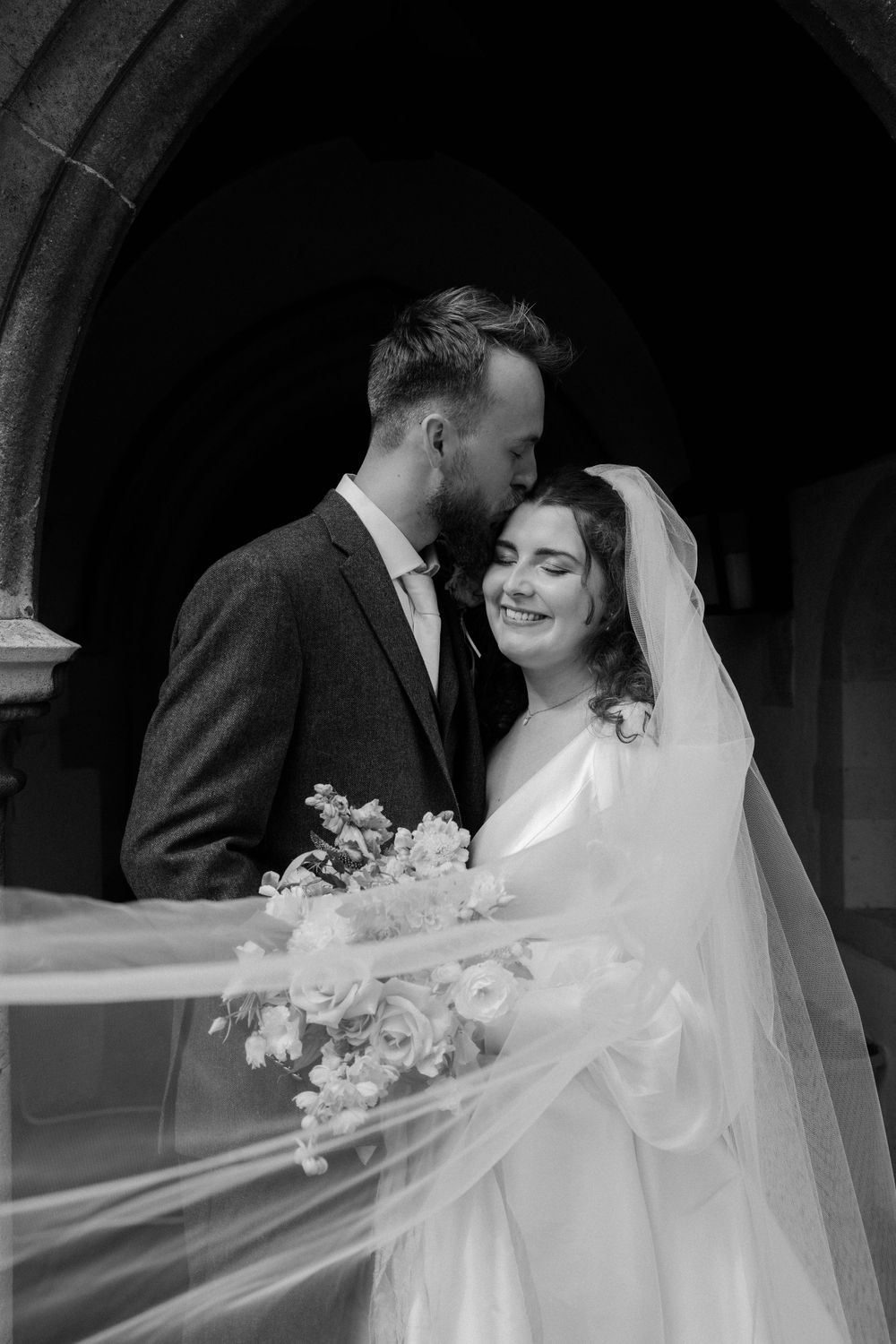 An intimate black and white wedding portrait captured under a stone church archway.