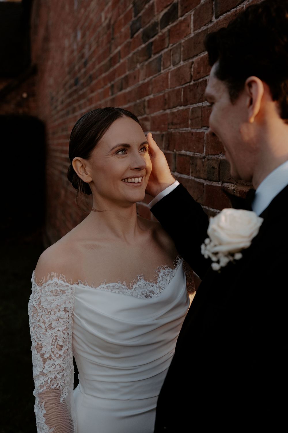 A groom tenderly touches his bride's face as she smiles warmly, both in wedding attire against a brick wall.