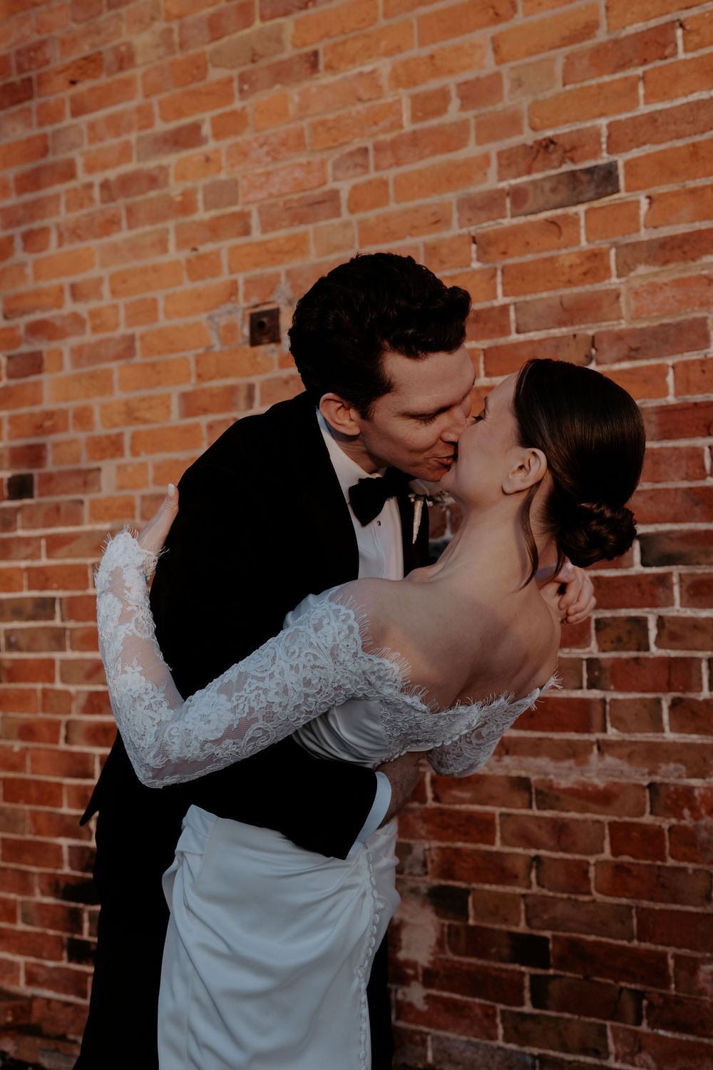 A bride and groom share a romantic kiss against a rustic red brick wall, the bride wearing a lace off-shoulder gown.
