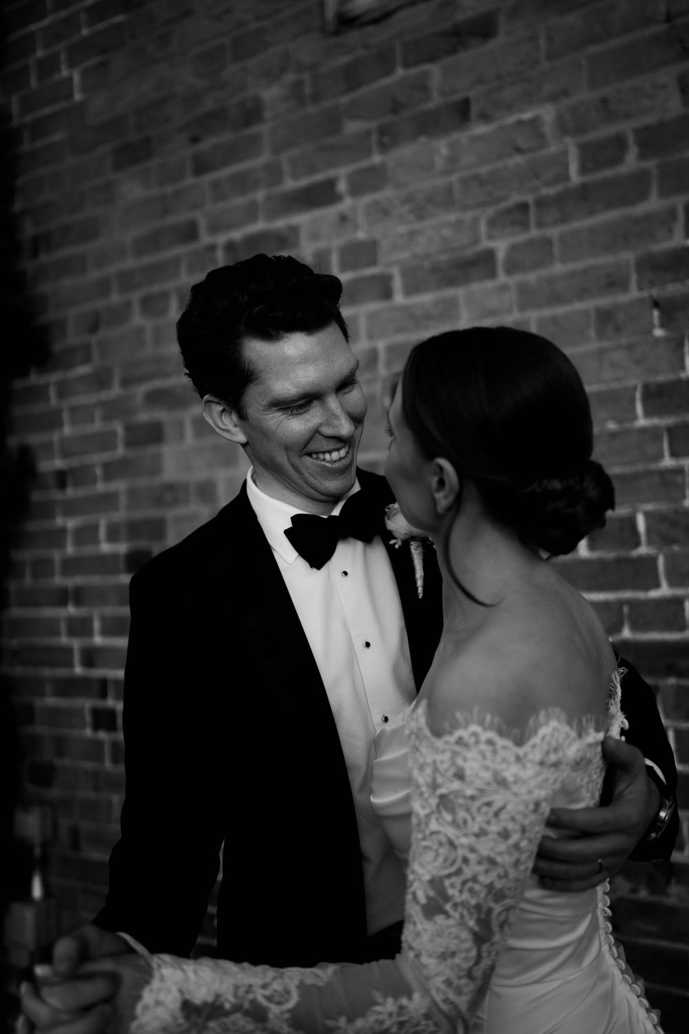 Black and white photo of a smiling groom in a tuxedo and bow tie embracing his bride in a lace wedding dress.
