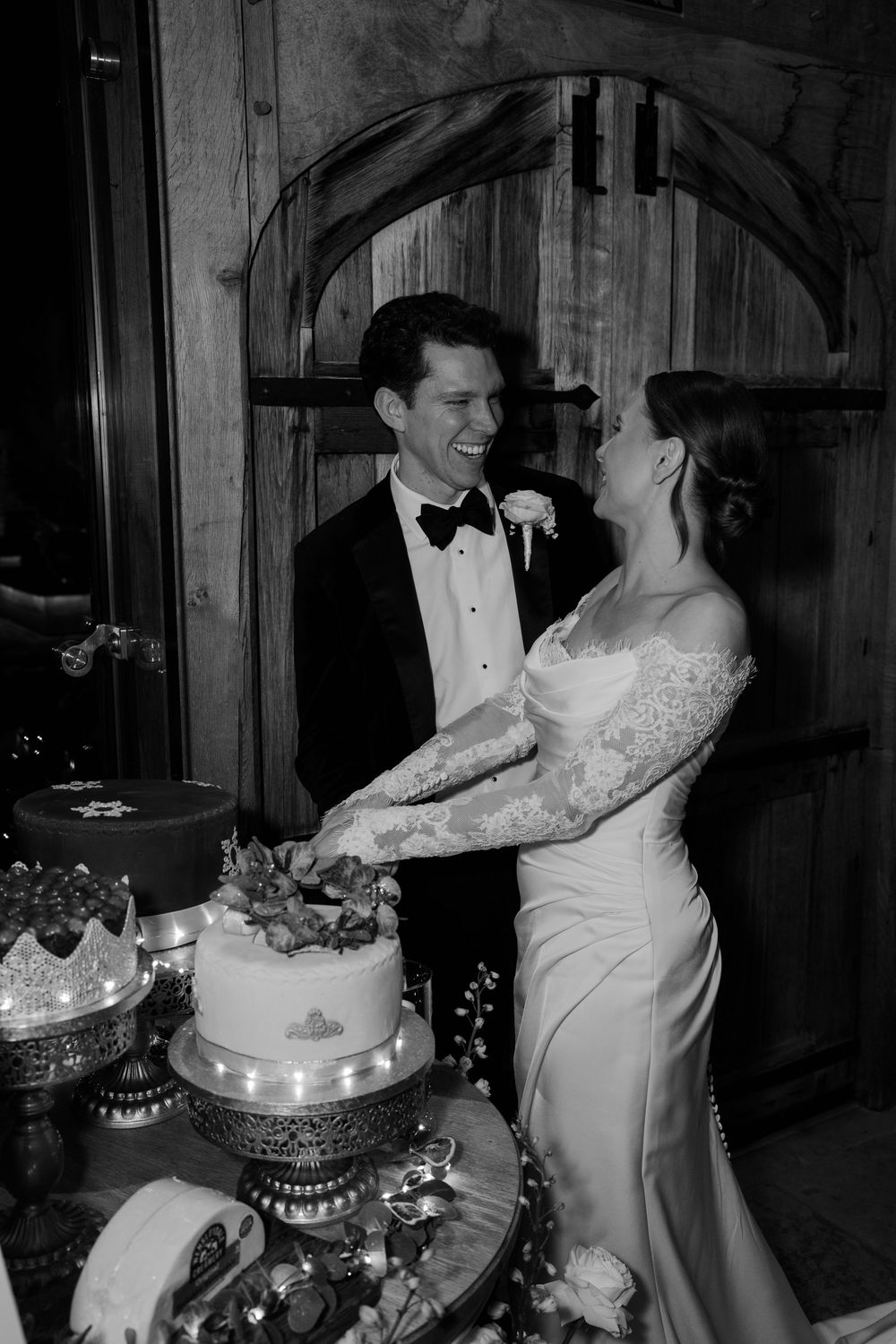 Black and white photo of smiling bride and groom cutting wedding cake together at elegant reception venue.