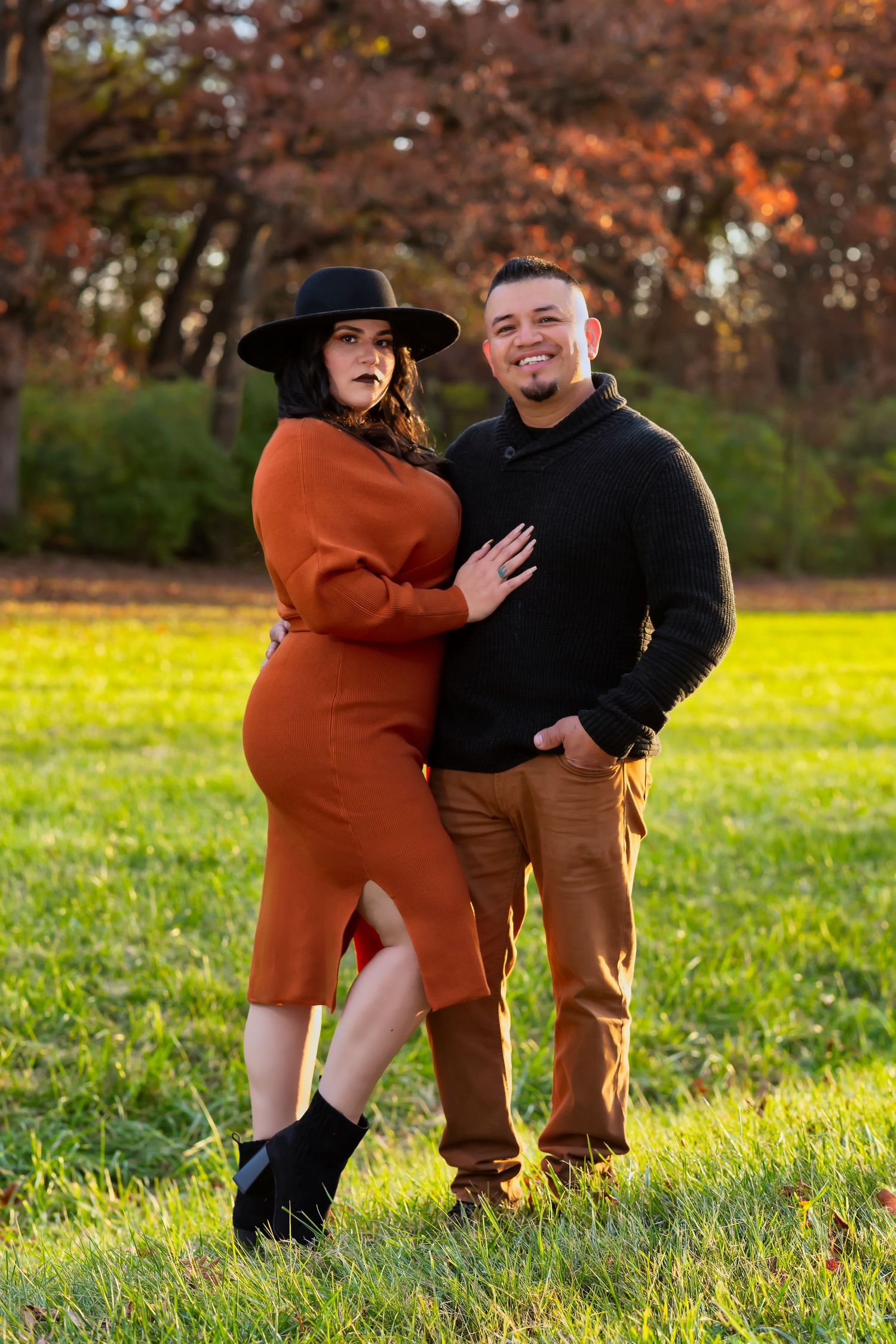 A couple poses outdoors in autumn, with her wearing a rust-colored dress and wide brim hat.