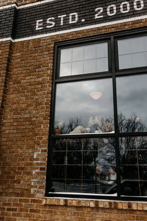 Brick building facade with 'ESTD. 2000' text and large industrial windows reflecting cloudy sky.