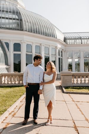 Elegant couple walking outside conservatory building with glass dome and ornate architecture.
