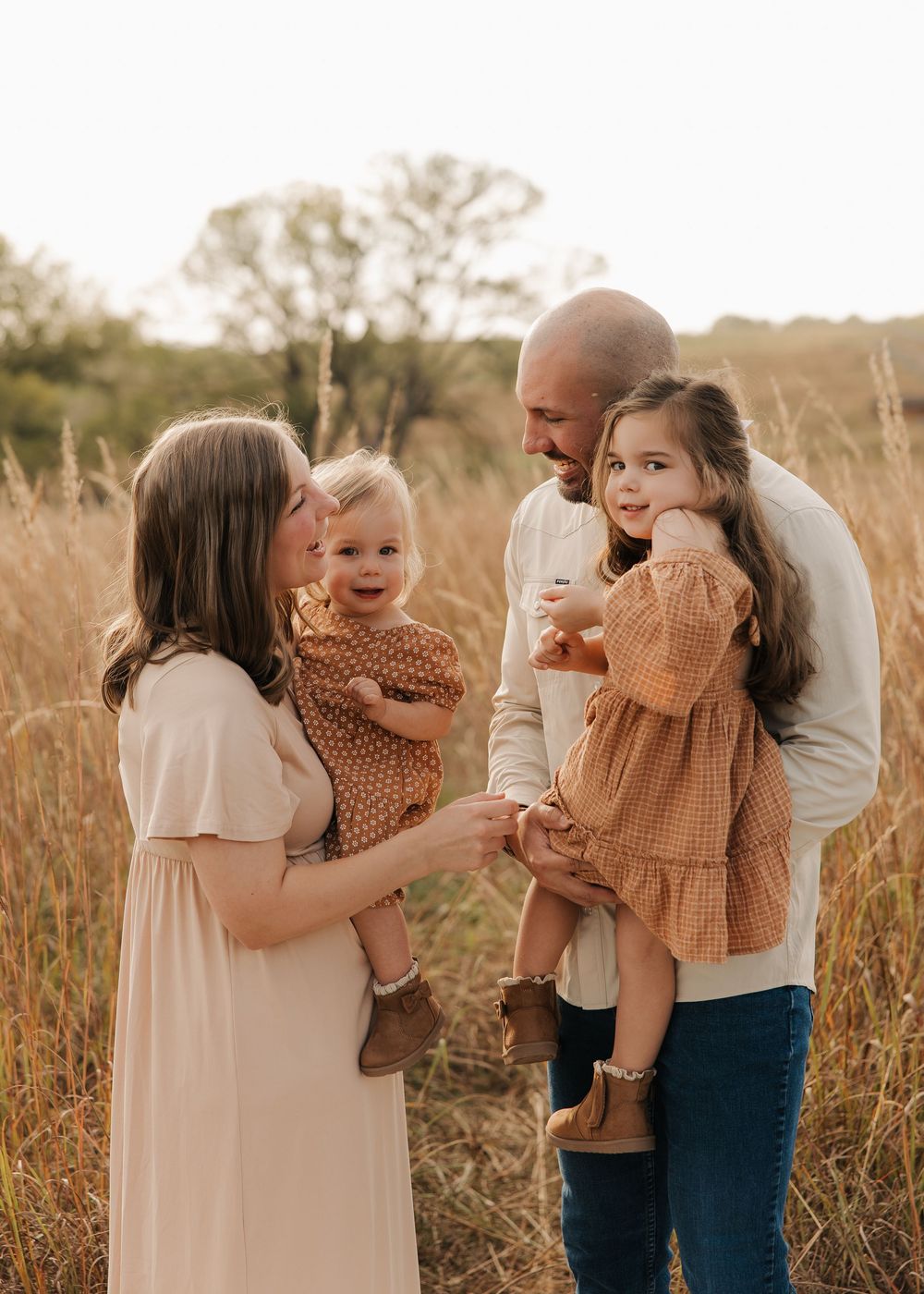 A family shares tender moments together in a golden wheat field at sunset wearing neutral toned clothing.