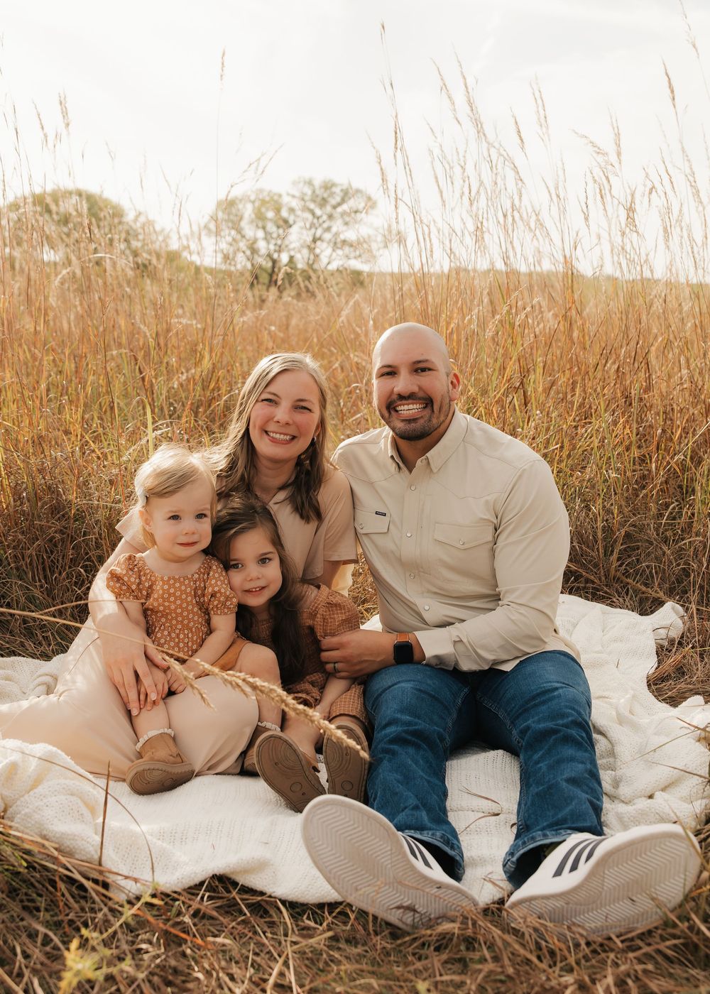 A family sits together on a white blanket in a field of tall golden grass during sunset.