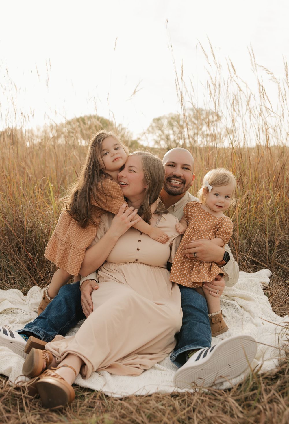 A family sits together on a white blanket in a golden field during sunset, sharing tender moments and embraces.