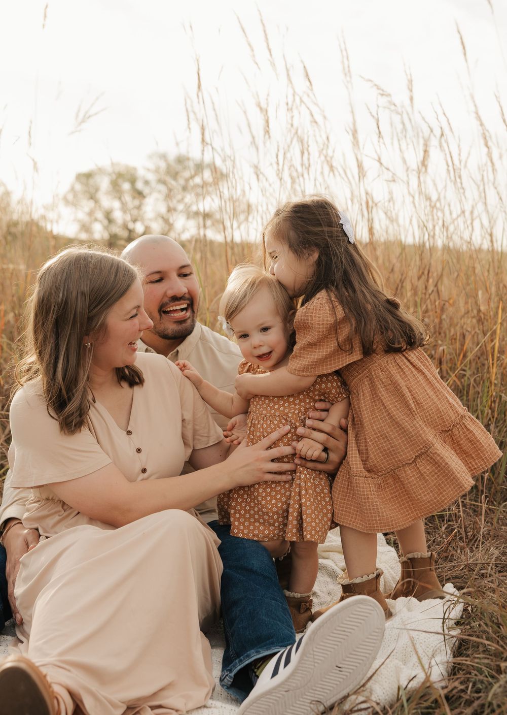A joyful family moment captured in warm tones as they embrace and laugh together in a field of tall grass at sunset.