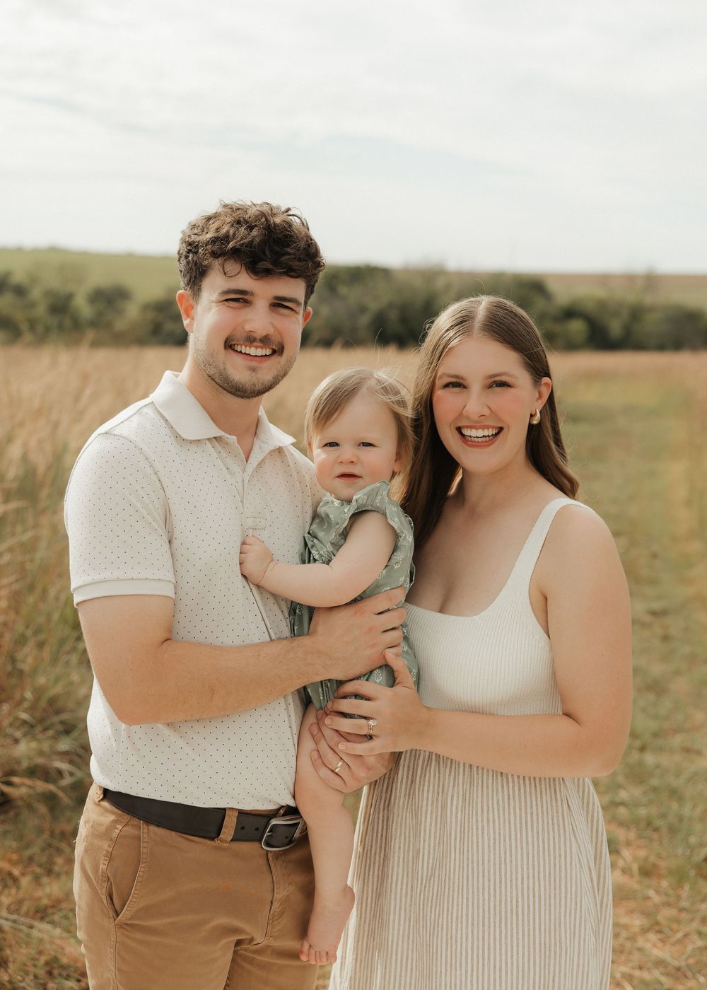 A family poses together in a grassy field wearing coordinated neutral tones during a sunny outdoor photoshoot.