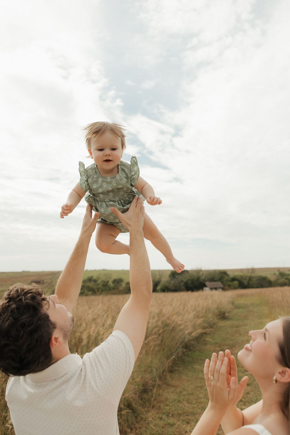 A parent lifts a small child wearing green clothing up into the air against a cloudy sky and grassy field background.