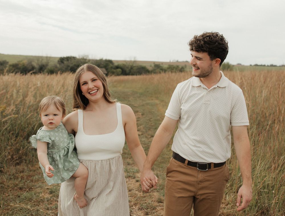 Family wearing neutral colored clothing walks together through a golden tall grass field at sunset.