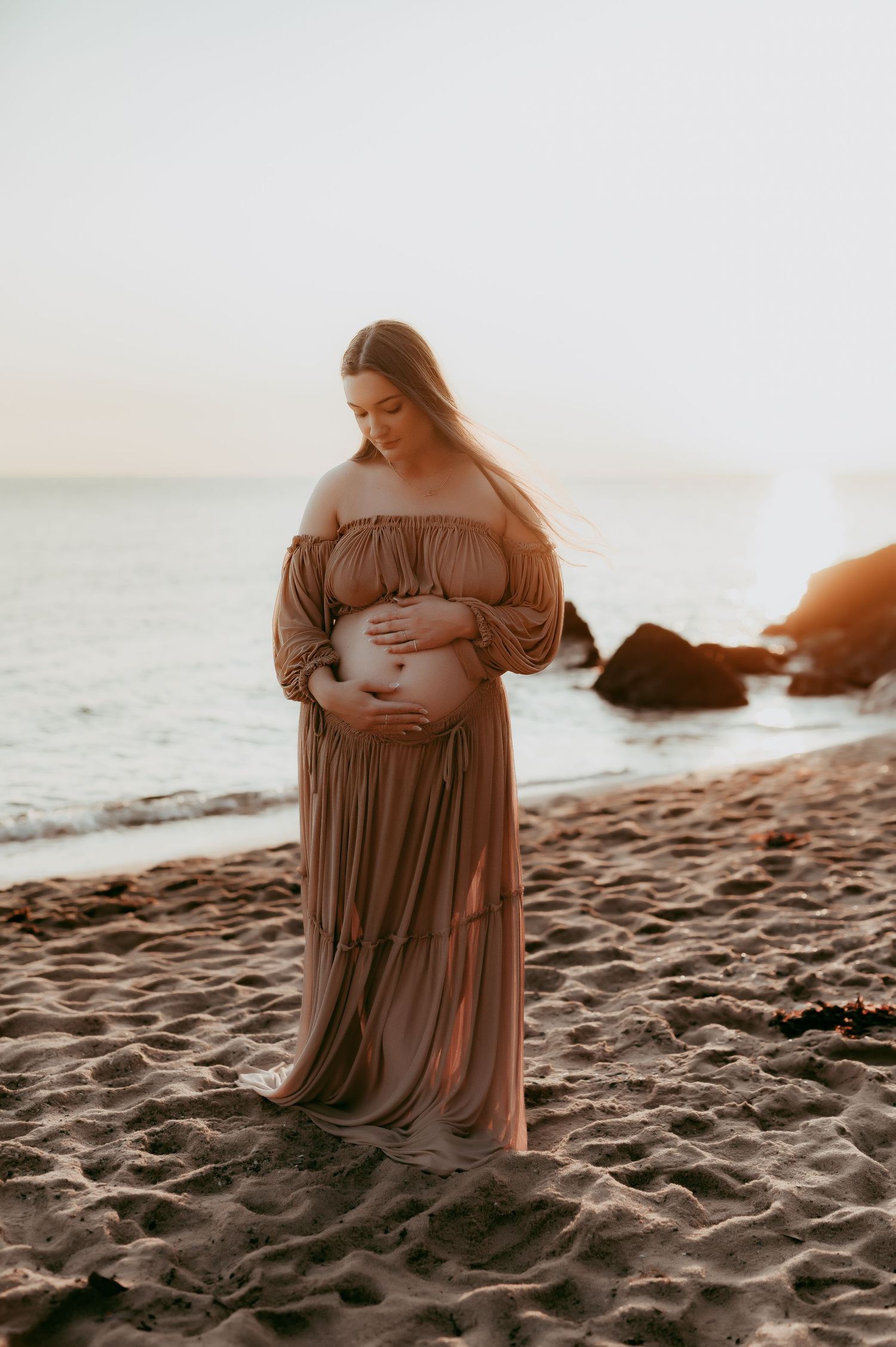 Ethereal maternity photo on beach at sunset with flowing brown dress and coastal rocks in background.