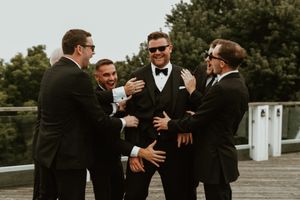 Group of groomsmen in black tuxedos sharing drinks and laughter outdoors.