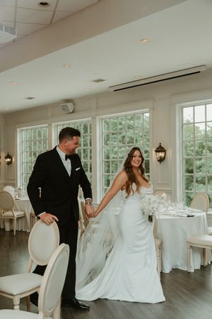 Couple dances playfully in an elegant white room with large windows and wedding reception decor.