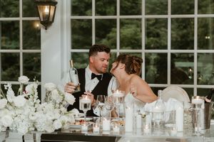 Elegant wedding reception table setting with white decor and candlelight near large windows.