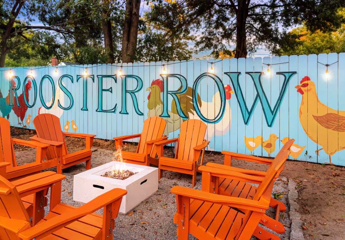 Orange Adirondack chairs arranged around a fire pit with a blue wall mural featuring 'Rooster Row' and chicken designs.