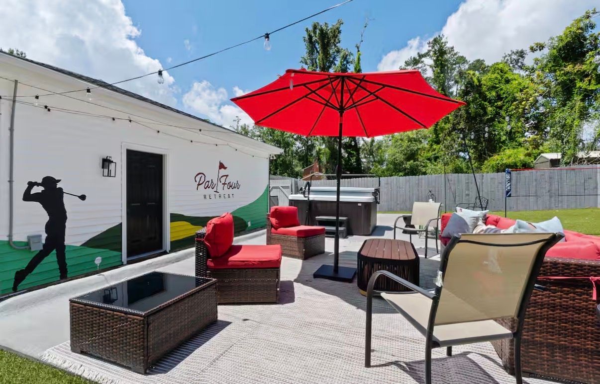 Outdoor patio area with red umbrella, wicker furniture, and golf-themed wall mural under bright blue skies. Par Four Retreat in Augusta, GA.