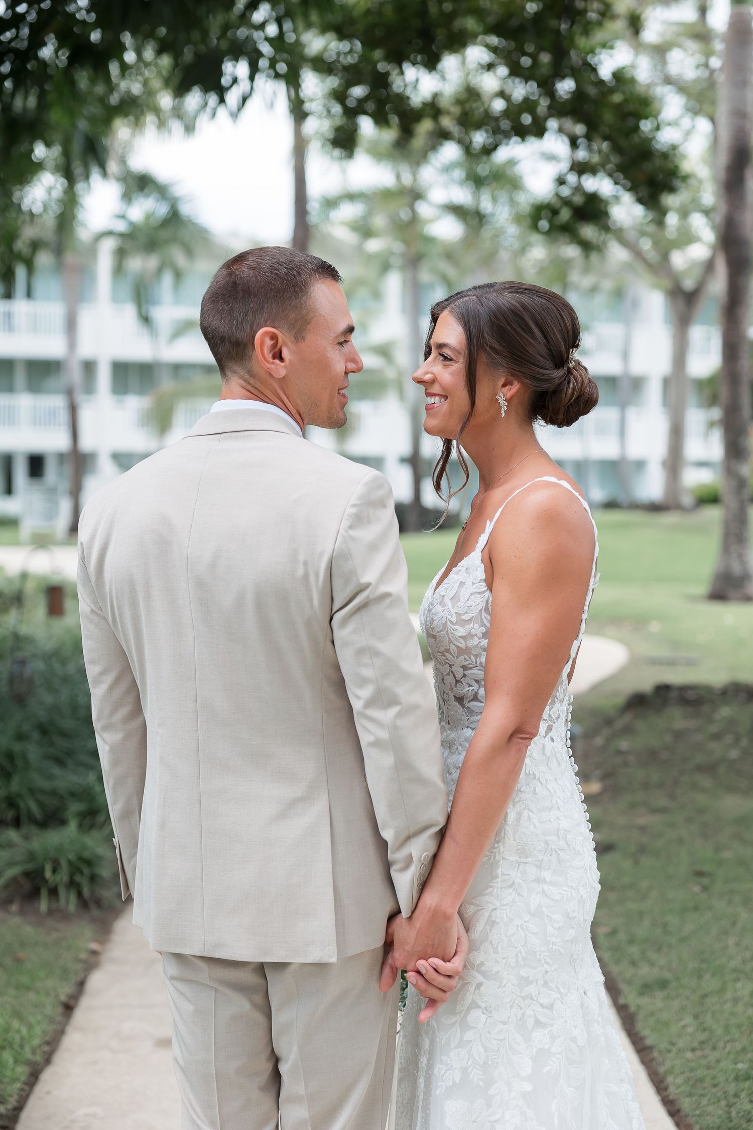 A bride and groom walk together on a grassy path surrounded by lush greenery.