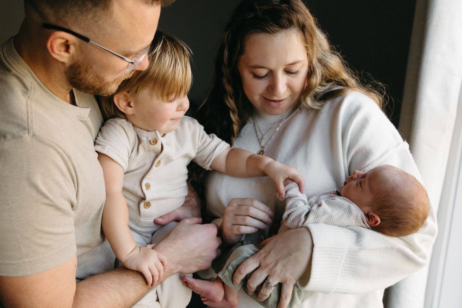 Mother holding her newborn baby while dad holds older sibling beside them touching newborn baby's hand.
