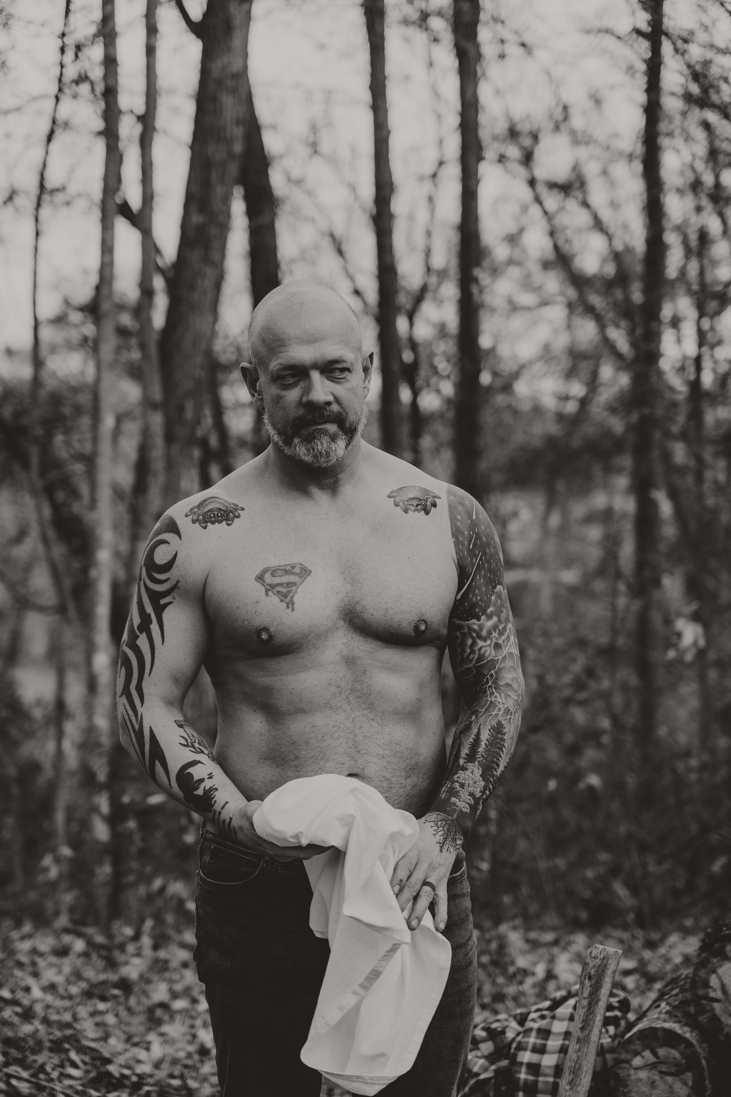 Artistic black and white photo of a shirtless tattooed person standing in a forest setting.