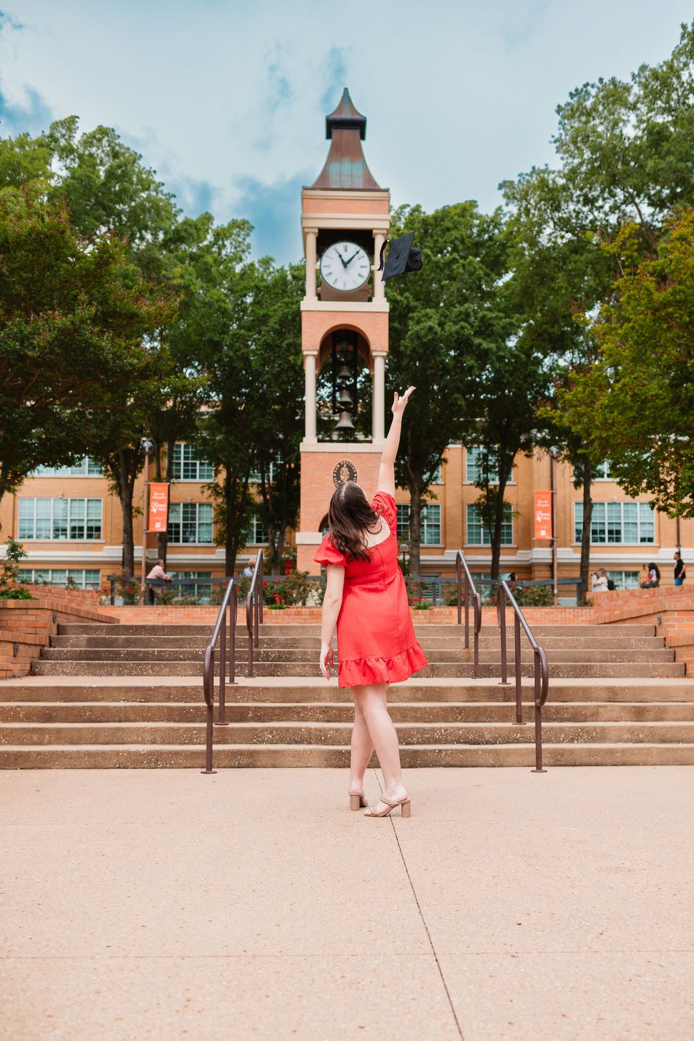 Graduate in red dress tossing cap in front of university clock tower on campus.