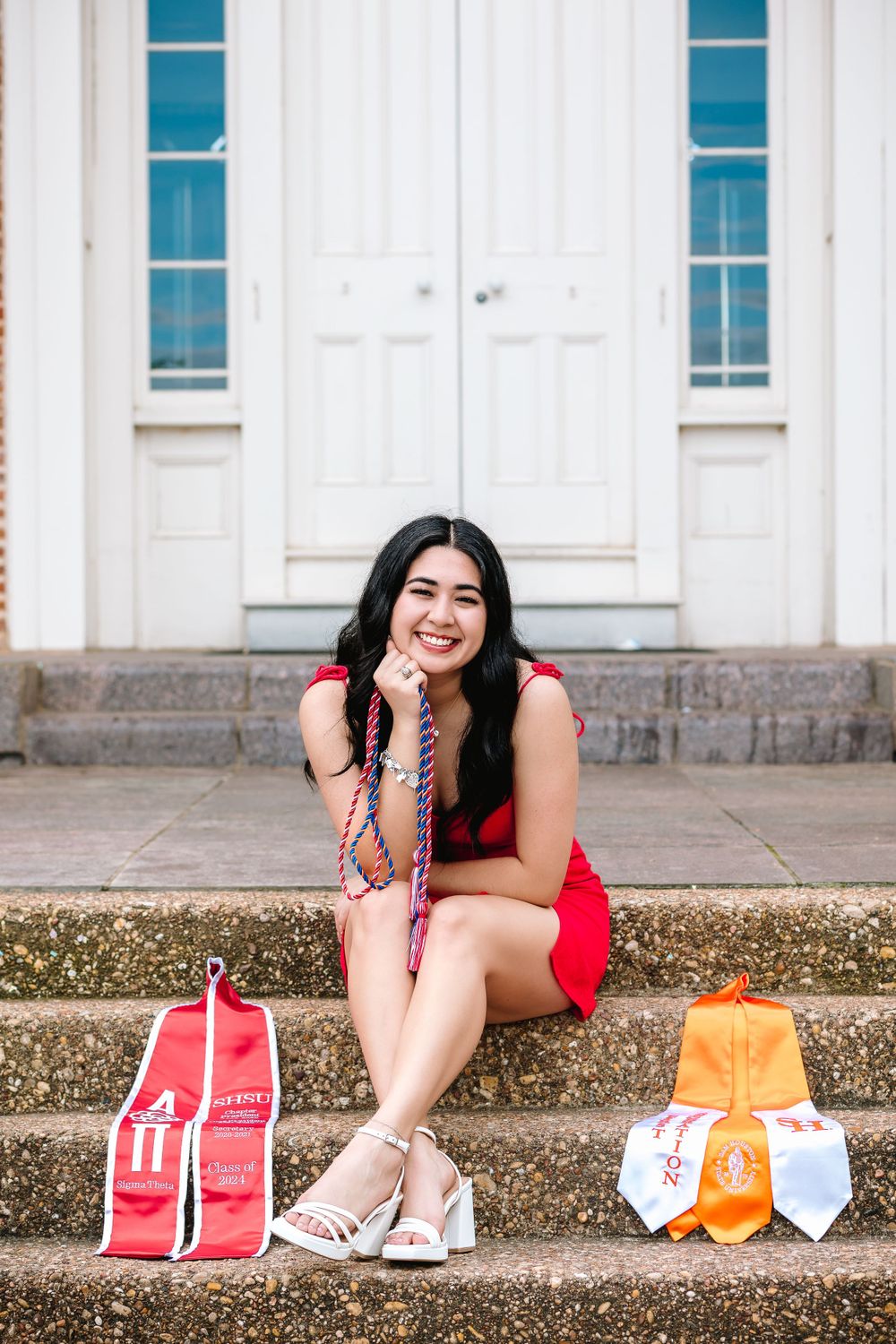 Smiling graduate in red dress sitting on steps with honor cords and graduation regalia.