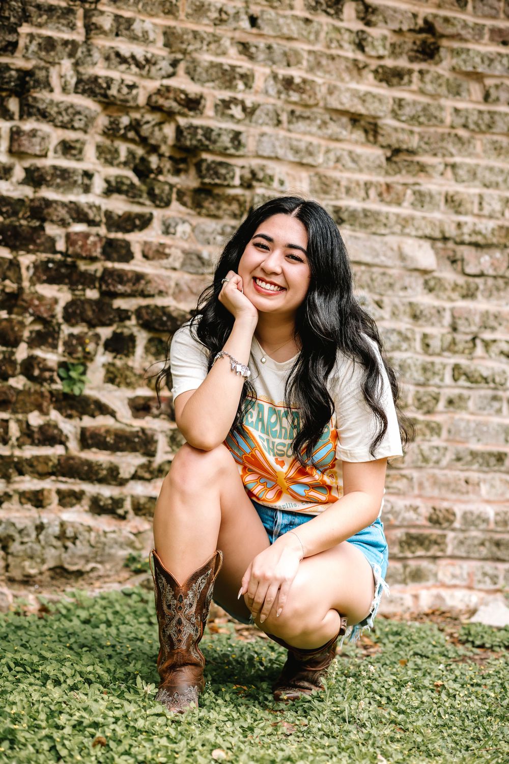Young woman in cowboy boots and denim shorts posing by rustic brick wall outdoors.