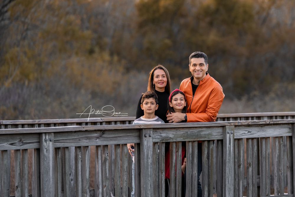 Family in orange and black outfits poses along wooden bridge railing during autumn sunset.