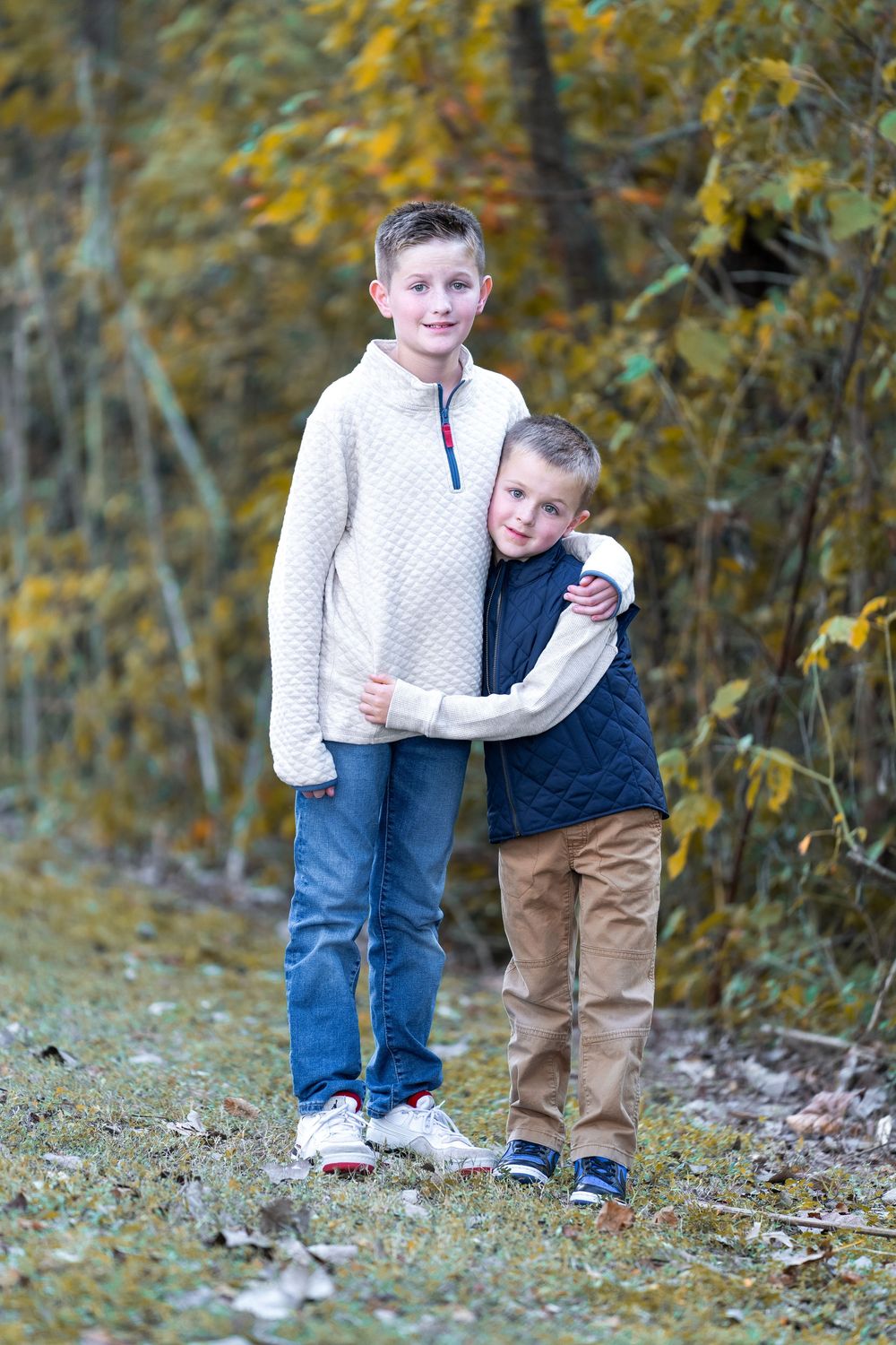 Two children in casual clothing play and embrace while standing outdoors among autumn foliage.