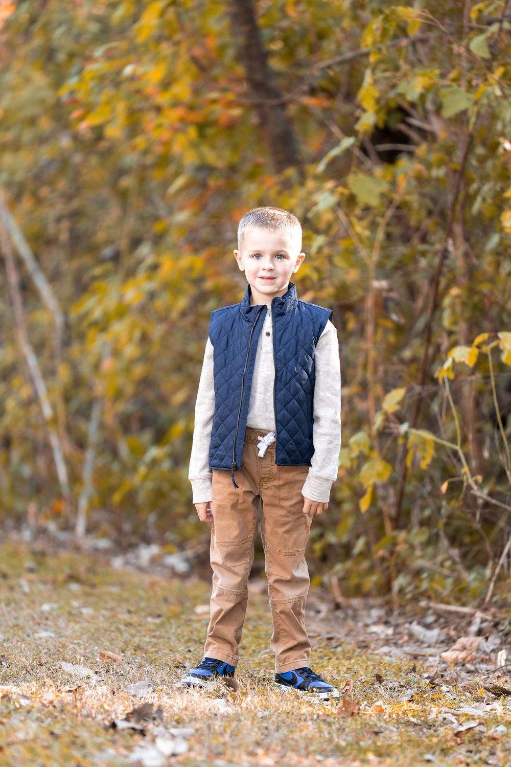A young child wearing a navy vest and brown pants stands among fallen autumn leaves in an outdoor setting.