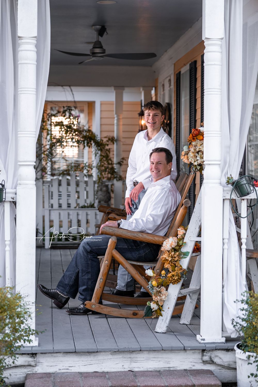 Two people relax in rocking chairs on a white-columned porch decorated with autumn garland.