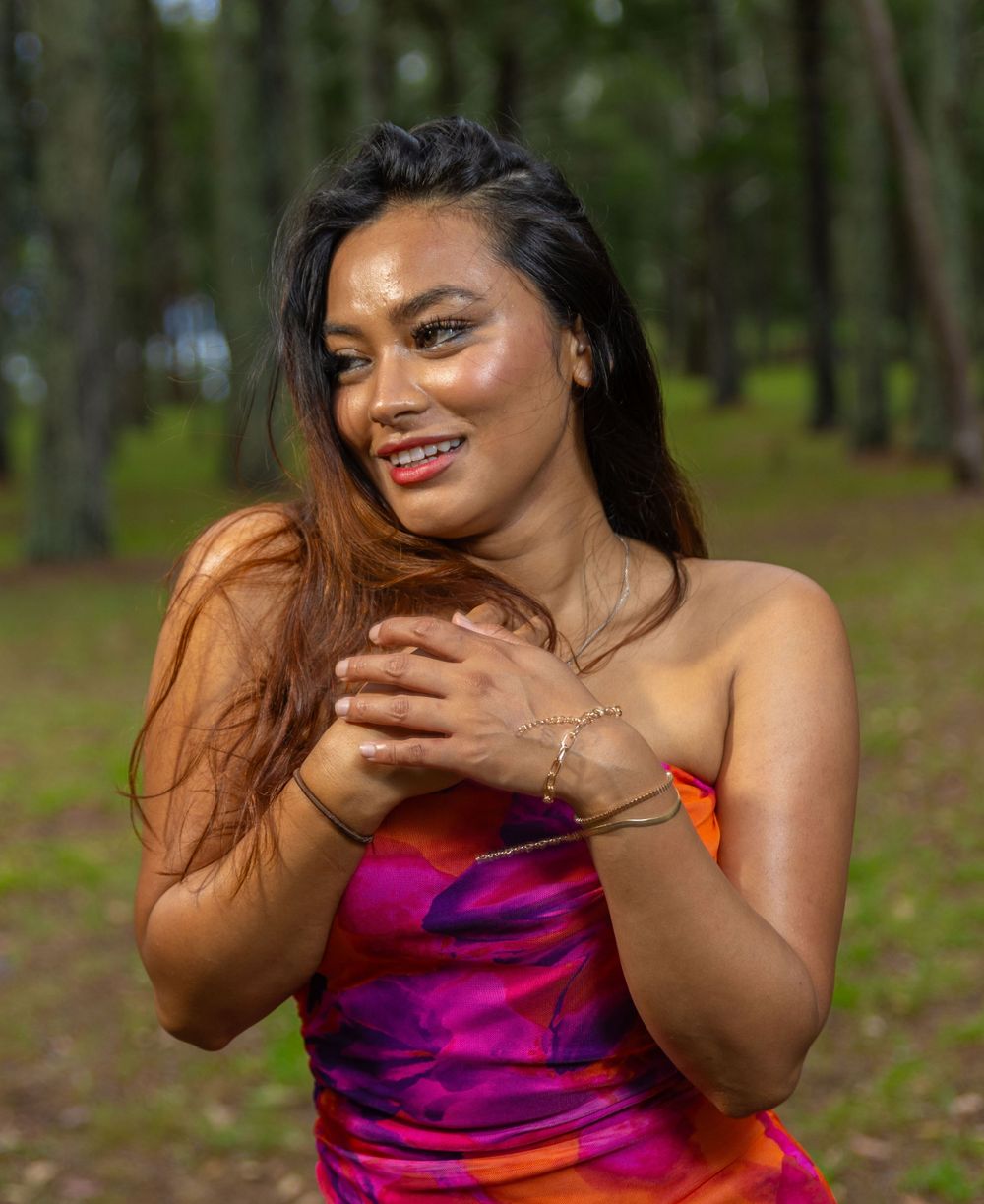 A woman wearing a colorful strapless dress smiles joyfully in a wooded outdoor setting.