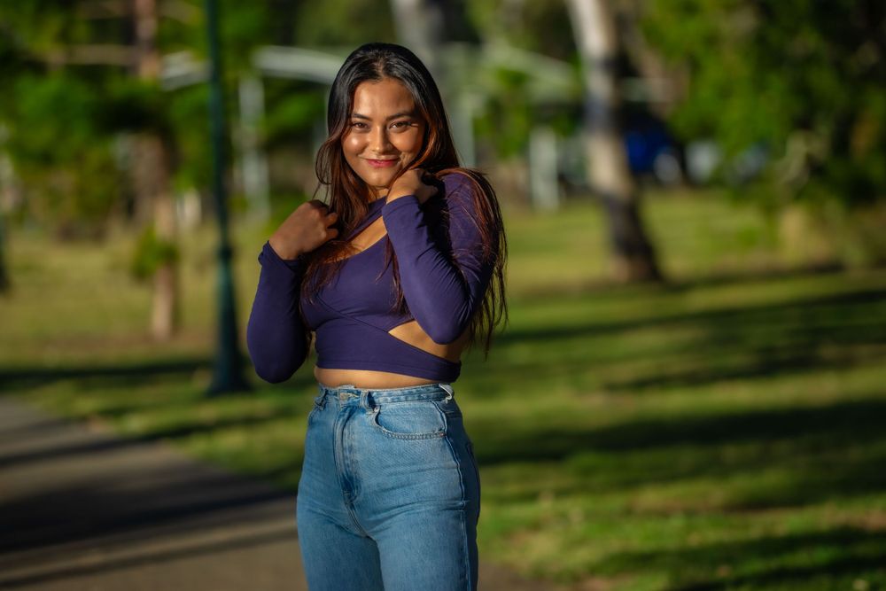 Woman in purple crop top and high-waisted jeans poses outdoors in a park setting.