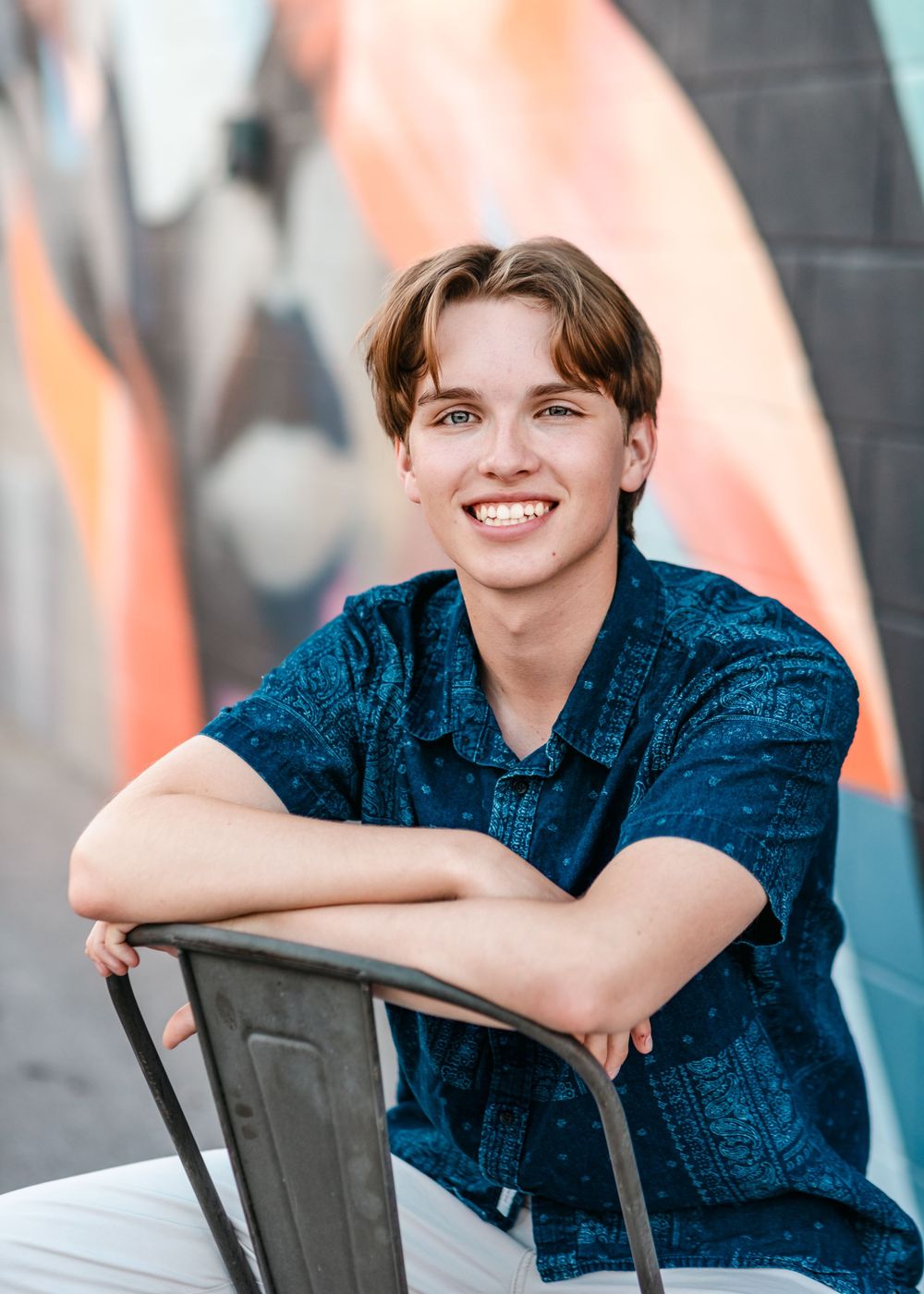 Young person in a blue patterned shirt sitting casually on a chair against a colorful graffiti wall.
