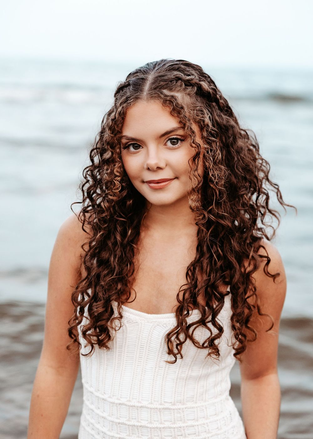 A person with long, naturally curly brown hair wearing a white tank top poses at the beach during golden hour.